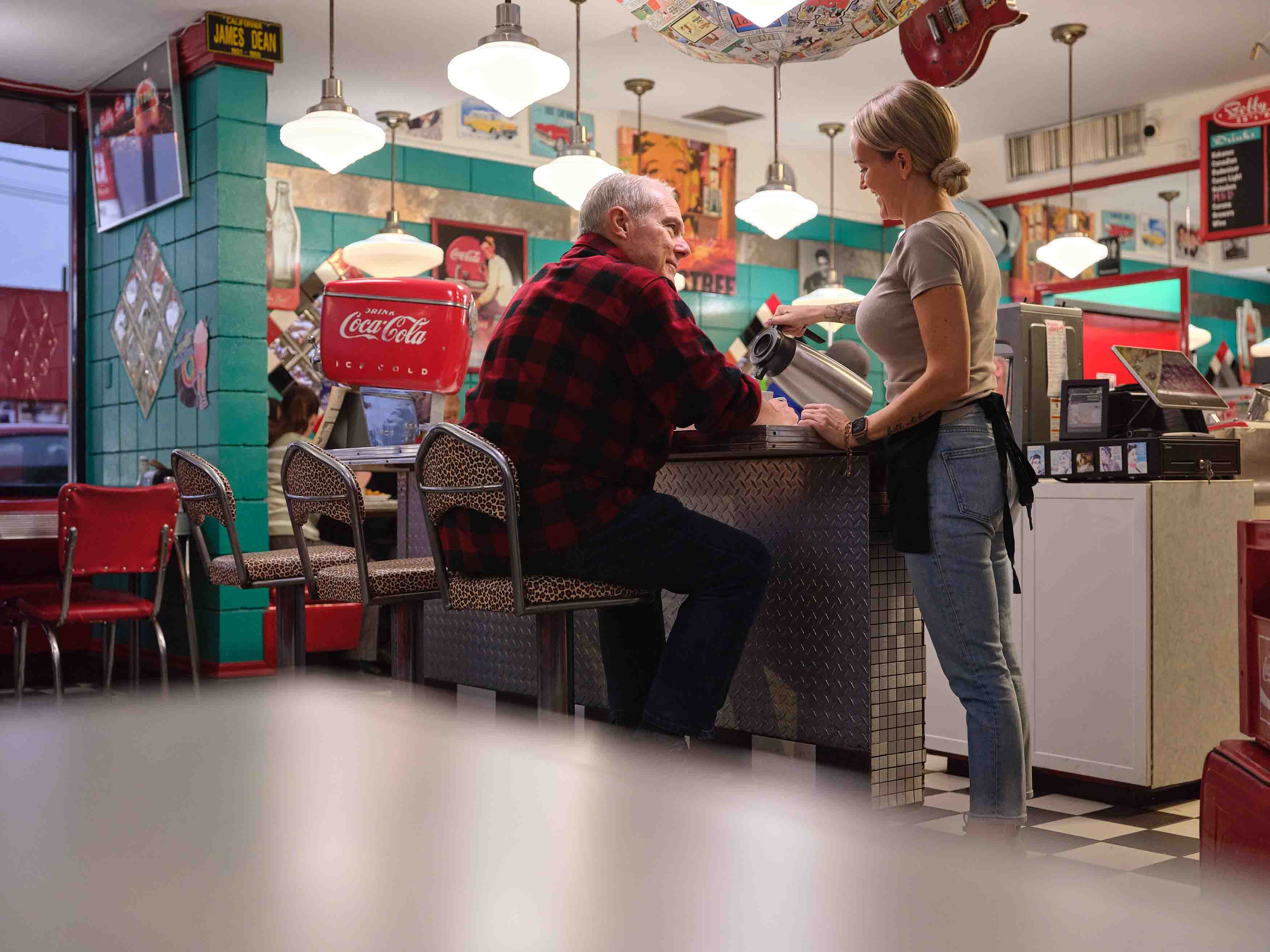 An older man sitting at the counter of a colourful diner while a woman stands beside him.