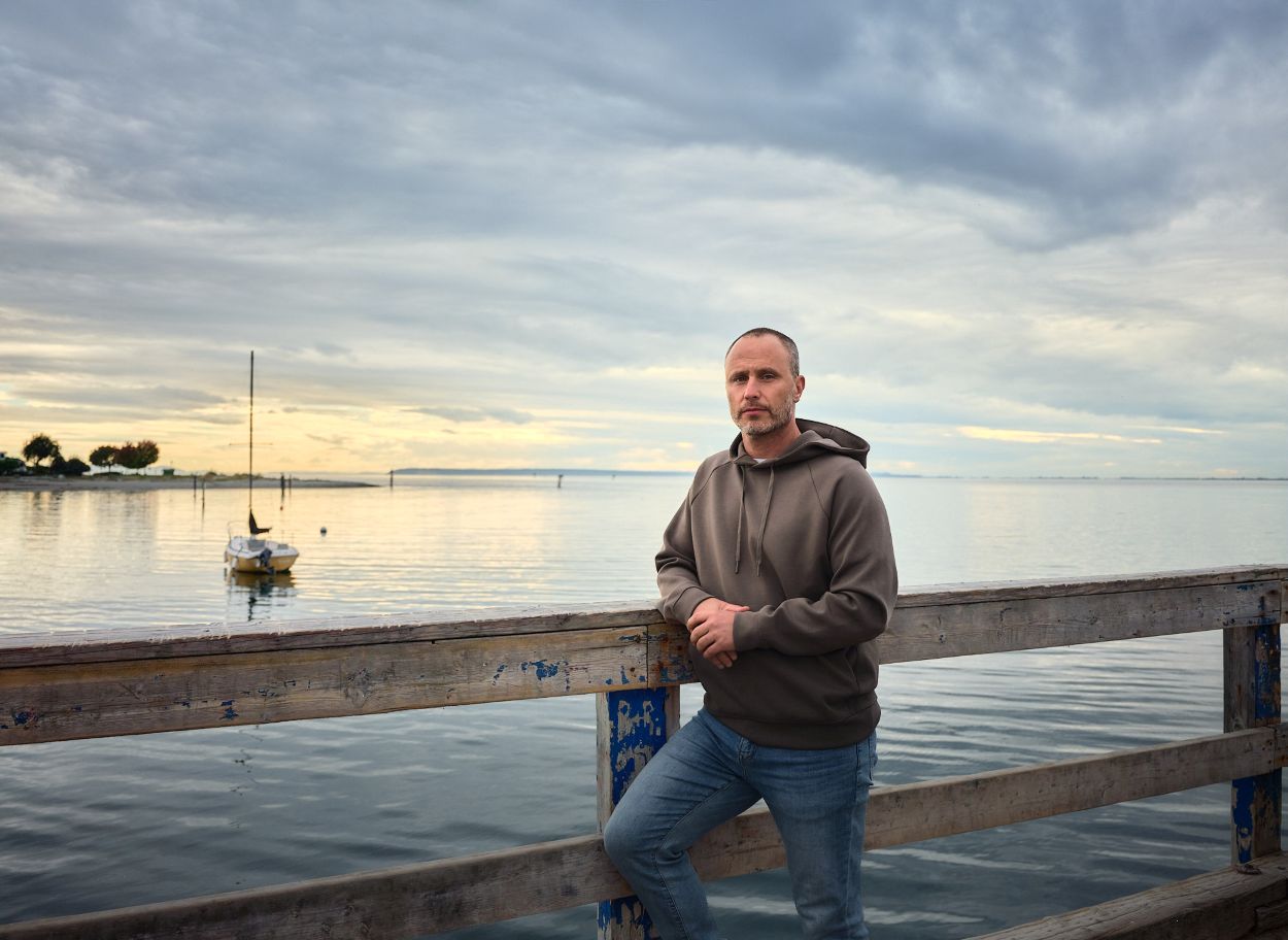 A man stands on a pier beside the ocean at sunset with a boat floating on the water. He has a serious look on his face.