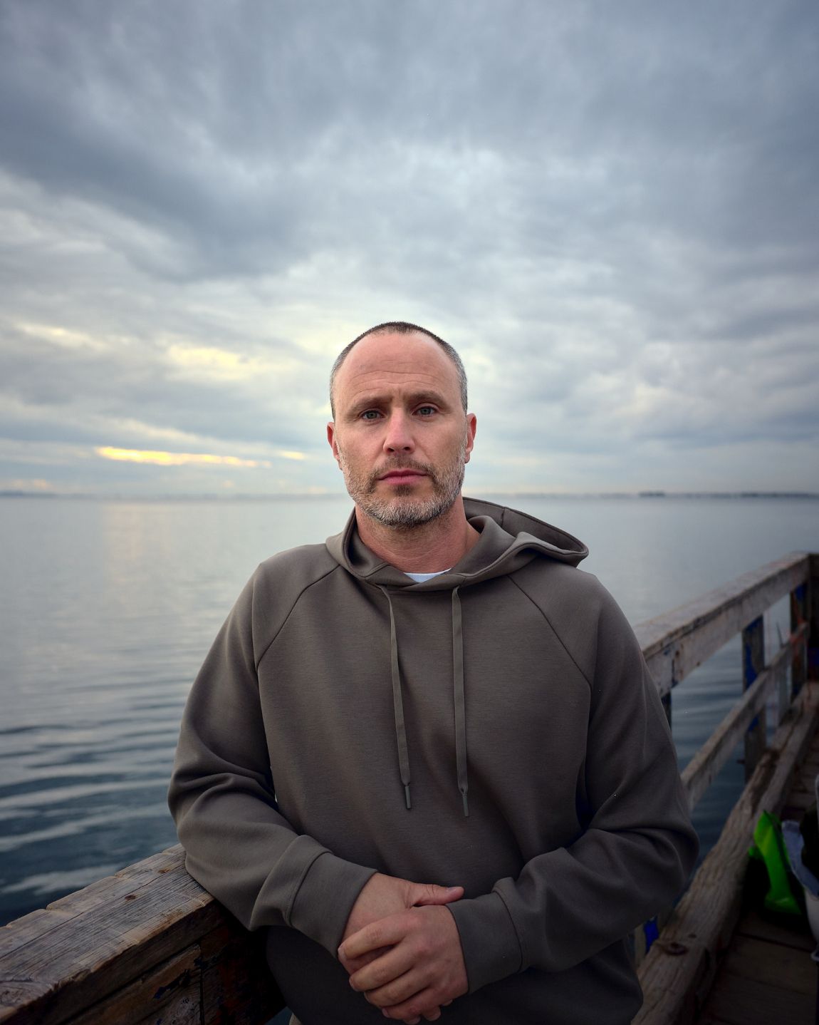 A man wearing a hoodie stands on a pier beside the water on an overcast day. He has a serious look on his face.
