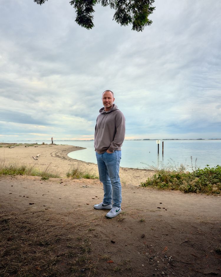 A man in casual attire stands on a beach with his hands in his jean pockets. There are clouds and ocean behind him.