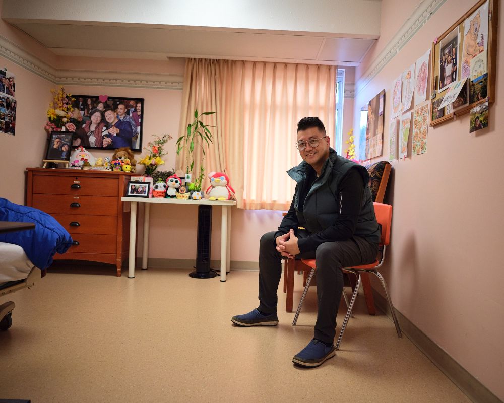 A man sits on a chair in his mother's long-term care room. Photos of the man and his mother hang in the background.