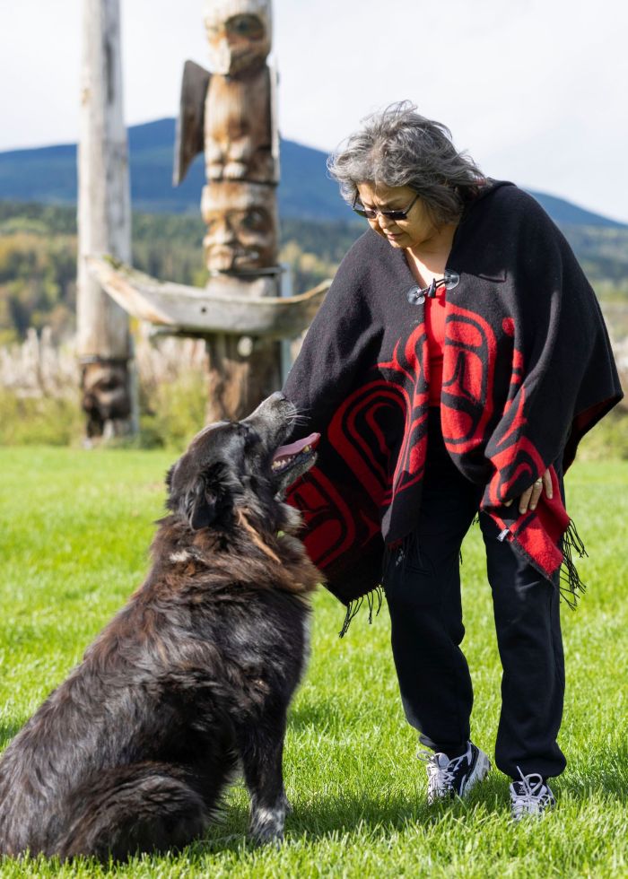 An Indigenous Elder stands in front of totem poles in the grass. She is petting a big black dog who looks back at her lovingly.