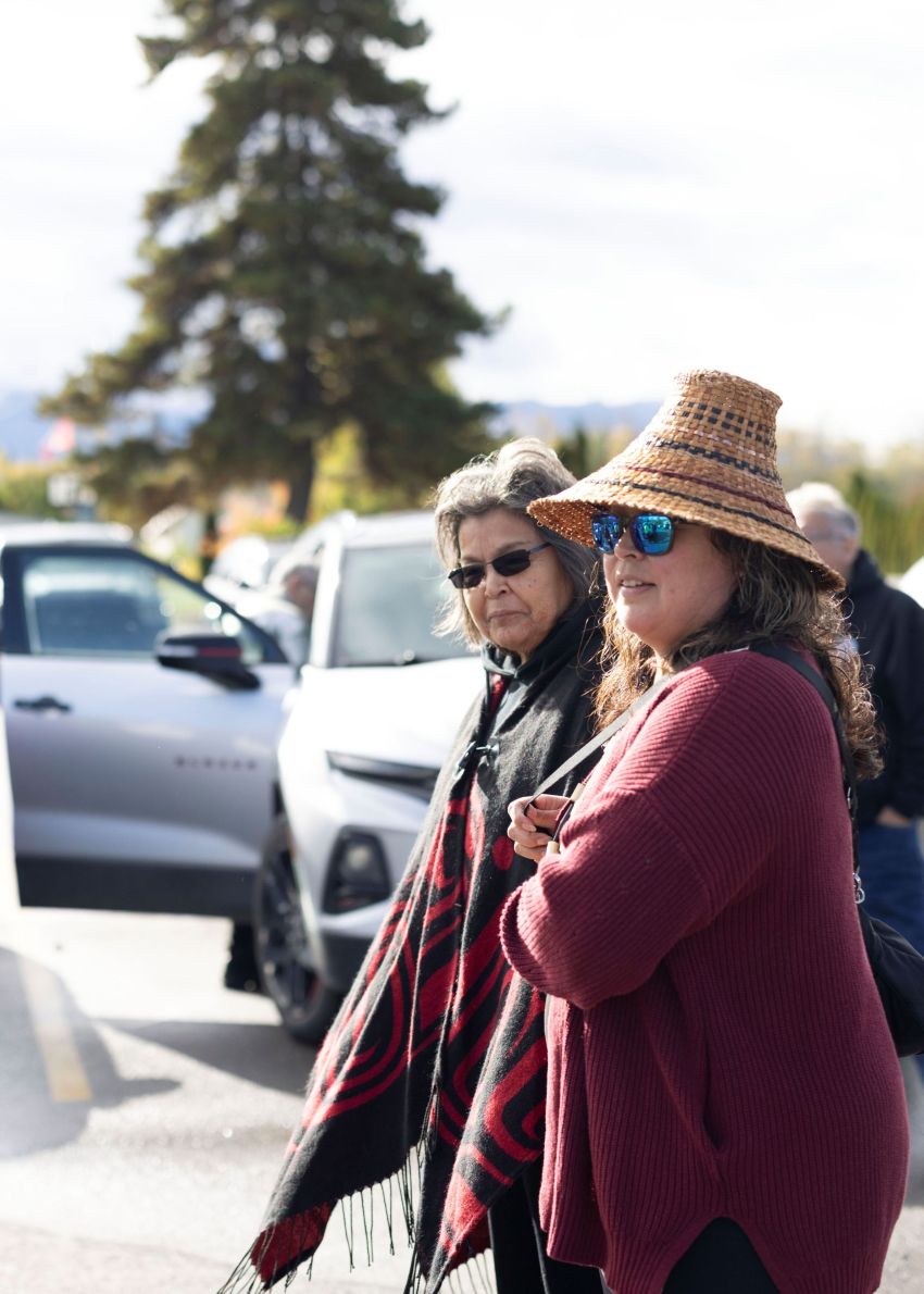 An Indigenous mother and daughter stand together outside. The daughter is wearing a woven hat.