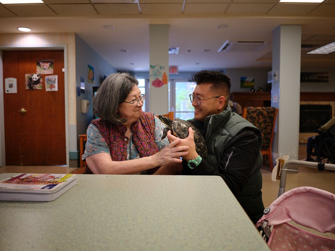 An elderly woman and her adult son sit at a table smiling. The son is holding a turtle.