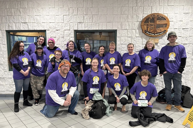 A large family poses in front of a white brick wall. They are smiling at the camera and all wearing purple shirts. 