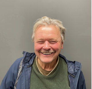 A man stands against a gray background, smiling for a headshot. He has gray hair and wears a green sweater.