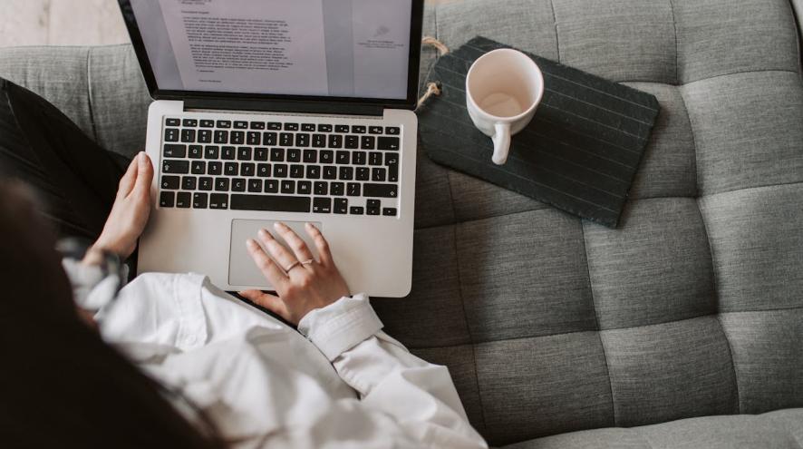 A person sits on the couch and uses a laptop to write a letter.