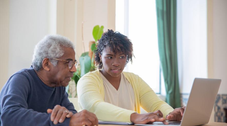 A young woman and an older man sit together at a table, both looking at a laptop screen. The woman is typing on the laptop while the man observes attentively.