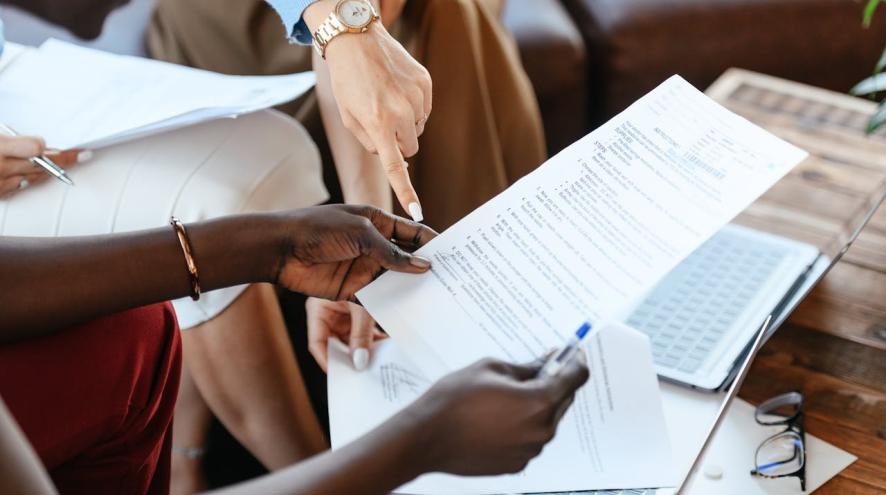 A close-up of hands holding and pointing at a document filled with text, with laptops open on a table in the background