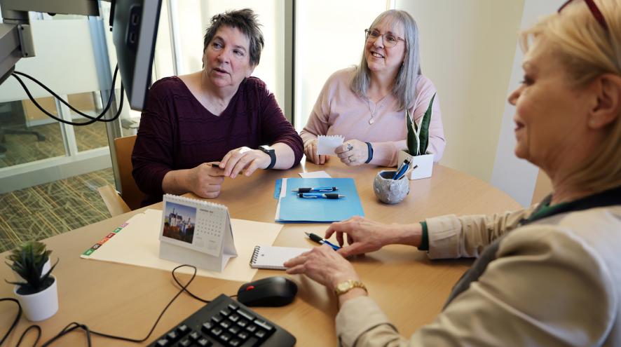 Two women sit in an office looking at a computer screen. A woman sits across the table from them, typing on a keyboard