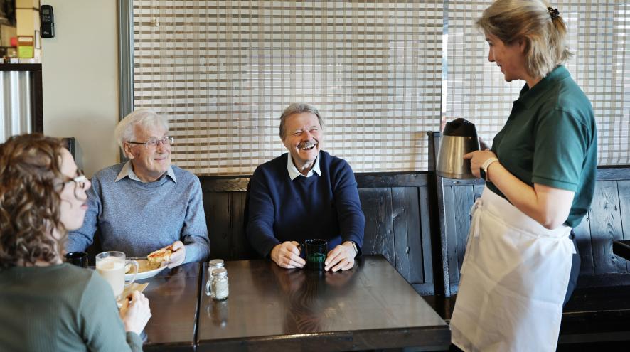 A server points to a coffee pot, standing near a table of three guests