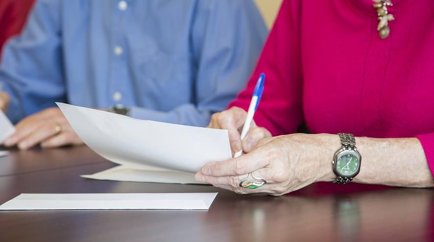 a close up of a man and woman writing letters at a wooden desk. You cannot see their faces.