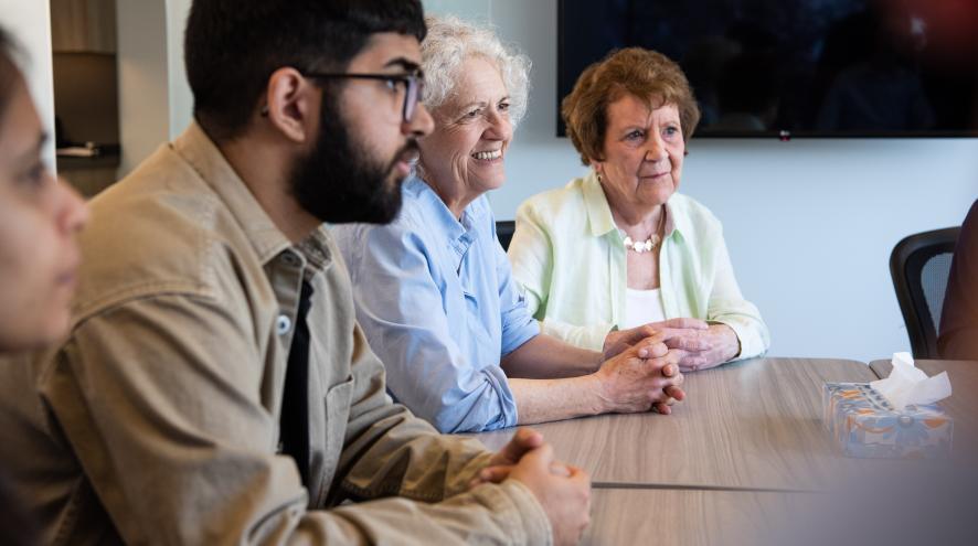 A group of people, diverse in age, race and gender, sit at a table. They talk to each other while smiling.