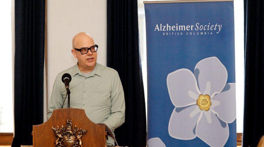A person speaks at podium within the BC Legislature's Hall of Honour, in front of an Alzheimer Society of BC banner.