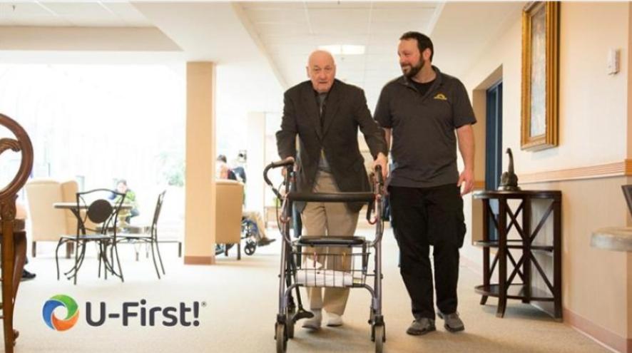 An elderly man using a walking frame walks alongside a male care aide