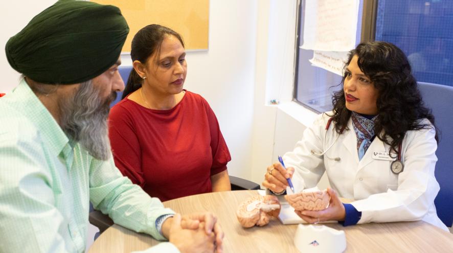 A health-care professional in a white coat speaks with a man wearing a green turban and a woman in a red shirt during a consultation.