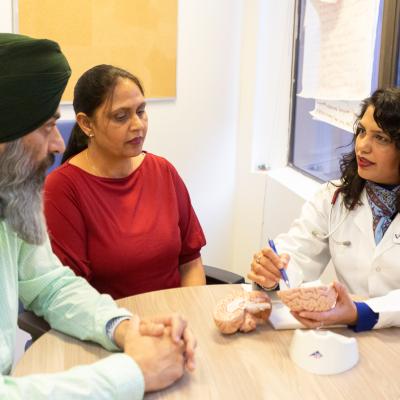 A health-care professional in a white coat speaks with a man wearing a green turban and a woman in a red shirt during a consultation.