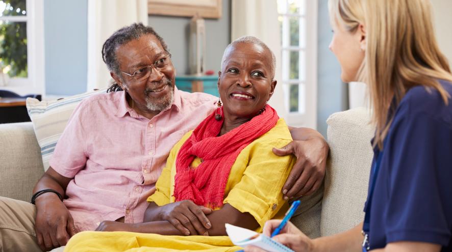 An Elderly Black woman wearing a yellow dress and red scarf, sitting on a couch with an elderly Black man in a pink button-up, who has his arm around her. They are speaking with a light-skinned blonde counsellor.