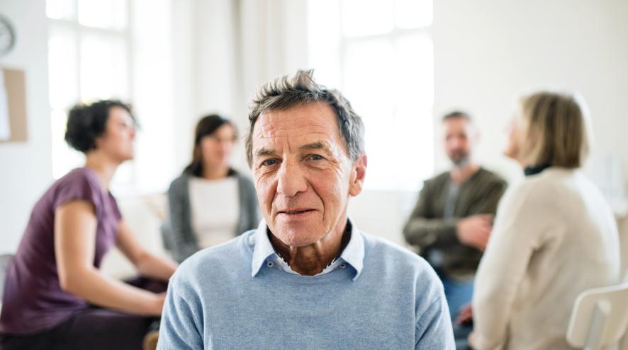 Senior man with light skin wearing a blue button-up shirt, with a group therapy session behind him, blurred in the background.