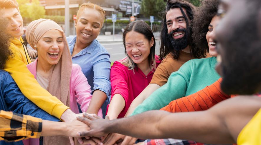 Group of people, diverse in race, gender, and age, wearing bright clothing and stacking hands in cooperation.