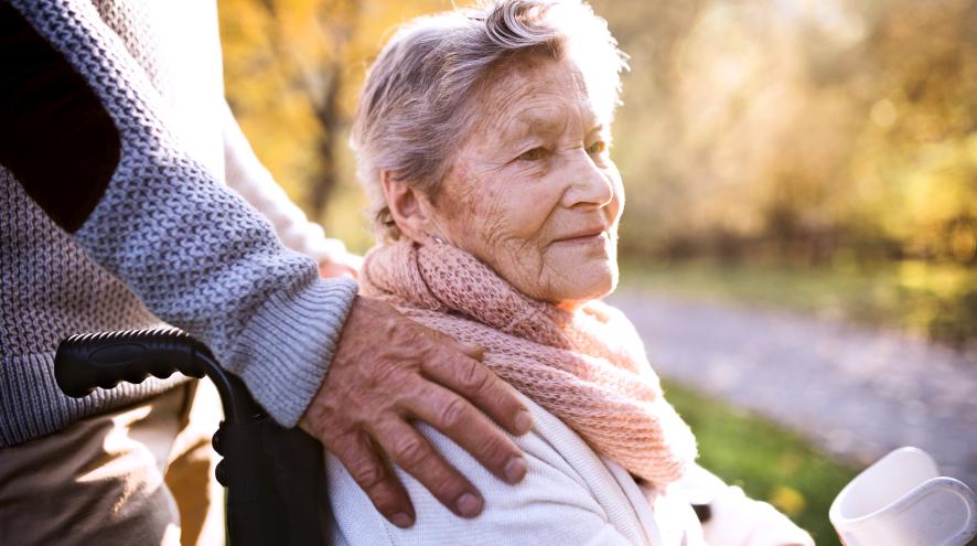 Senior woman with pale skin and short grey hair, sitting in a wheelchair outdoors, as a person out of frame rests a hand on her shoulder.