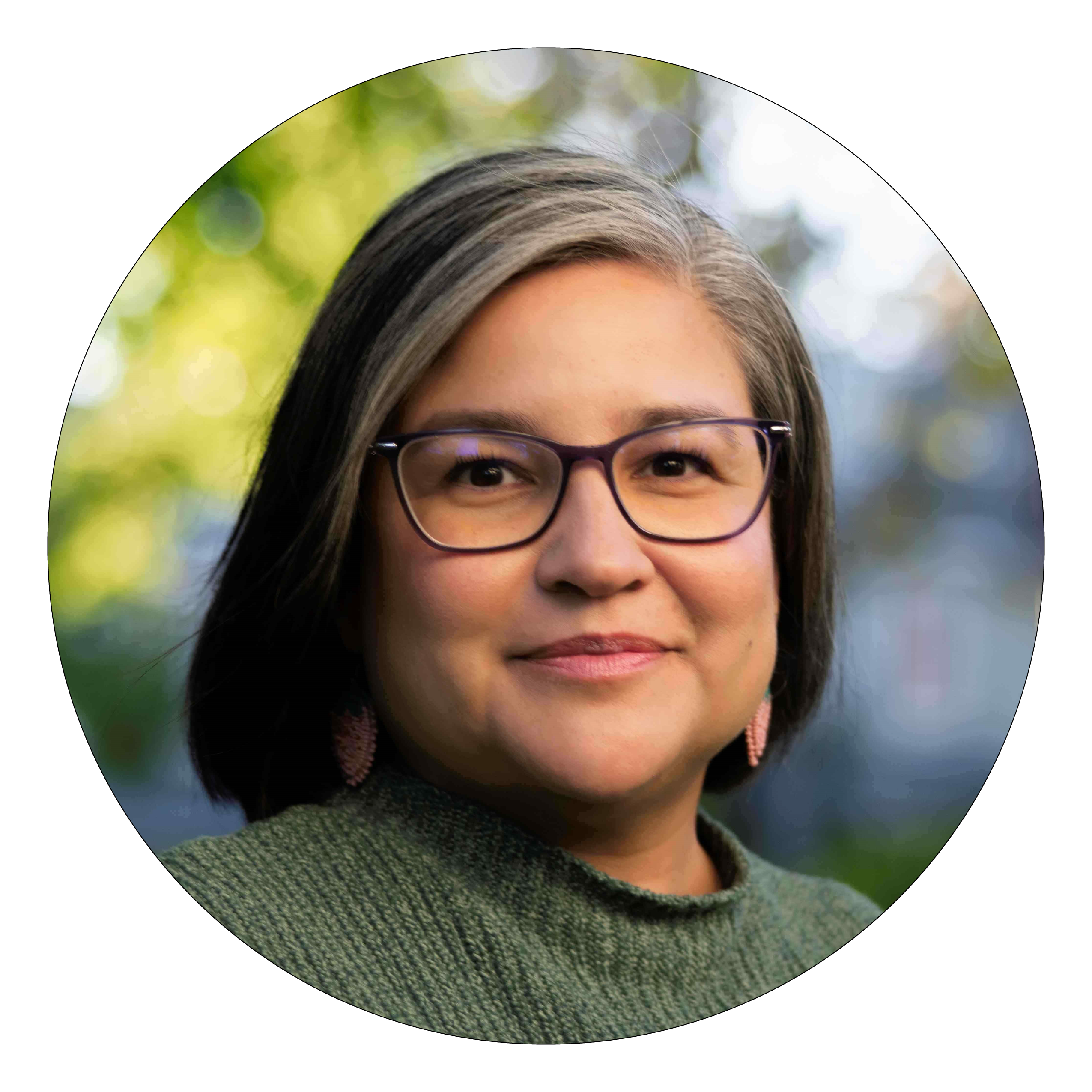 A woman with black and grey hair, green sweater and black frame glasses smiles