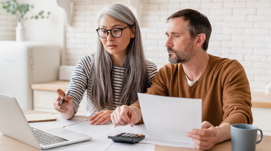 A midlife woman and man sit at a kitchen counter with laptop, calculator, pens and papers