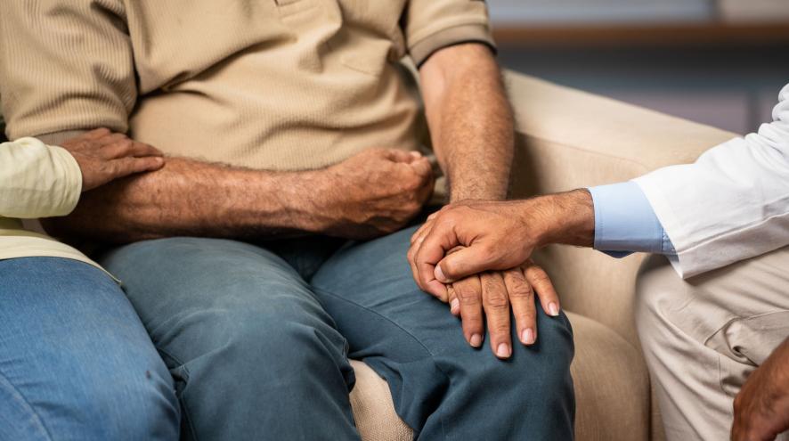 A person in a white medical jacket holds the hand of a person in blue pants and beige sweater