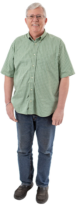 A man in a green button-up shirt, jeans, and glasses is smiling. 