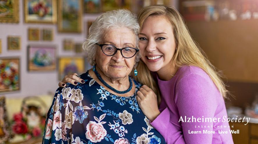 A young woman standing next to her grandmother in front of a picture wall in her home.